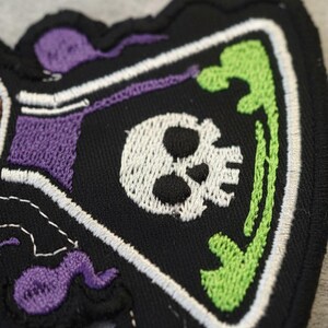 May include: Embroidered patch with a skull design in purple, green, and white. The patch is black with a white skull, green accents, and purple details. The patch has a unique shape and is made of fabric.