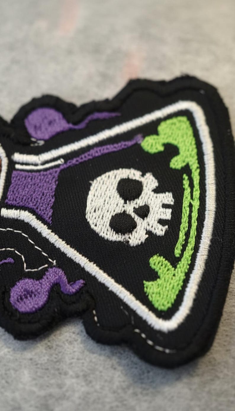 May include: Embroidered patch with a skull design in purple, green, and white. The patch is black with a white skull, green accents, and purple details. The patch has a unique shape and is made of fabric.