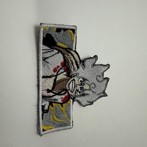 May include: A rectangular embroidered patch featuring a stylized anime character with spiky light gray hair, a wide grin, and red accents on a white garment. The background of the patch is gray with yellow swirling patterns and a white border.