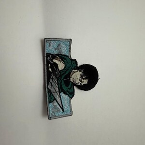 May include: A rectangular embroidered patch featuring a character with black hair wearing a dark green cloak and holding a silver blade. The character is set against a light blue background with white cloud-like patterns, all bordered by a black stitched edge.