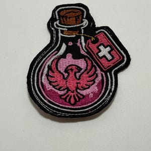Peut inclure: Un écusson brodé en forme de flacon rond rempli de liquide rose. À l'intérieur du flacon se trouve un emblème de phénix rose. Le flacon possède un bouchon en liège marron et une petite étiquette rectangulaire rose avec une croix blanche.