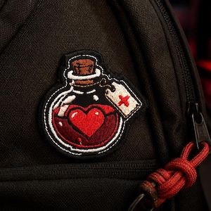 May include: Embroidered patch featuring a potion bottle with a red heart inside. The bottle is clear with a brown cork and a white tag with a red cross. The patch is attached to a black backpack.