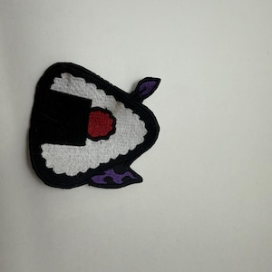 Op de afbeelding: Een geborduurde patch in de vorm van een driehoekige rijstbal, of onigiri, met witte rijst, een zwarte zeewierwikkel, een rood midden en paarse vlamachtige accenten aan de boven- en onderkant, alles omlijnd met een dikke zwarte rand.