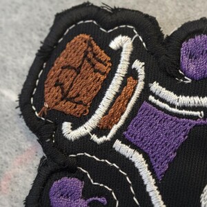 May include: Embroidered patch with a black background and a detailed design featuring brown, purple, and white stitching. The patch has a textured appearance with a stitched border, showcasing a unique, handcrafted aesthetic.