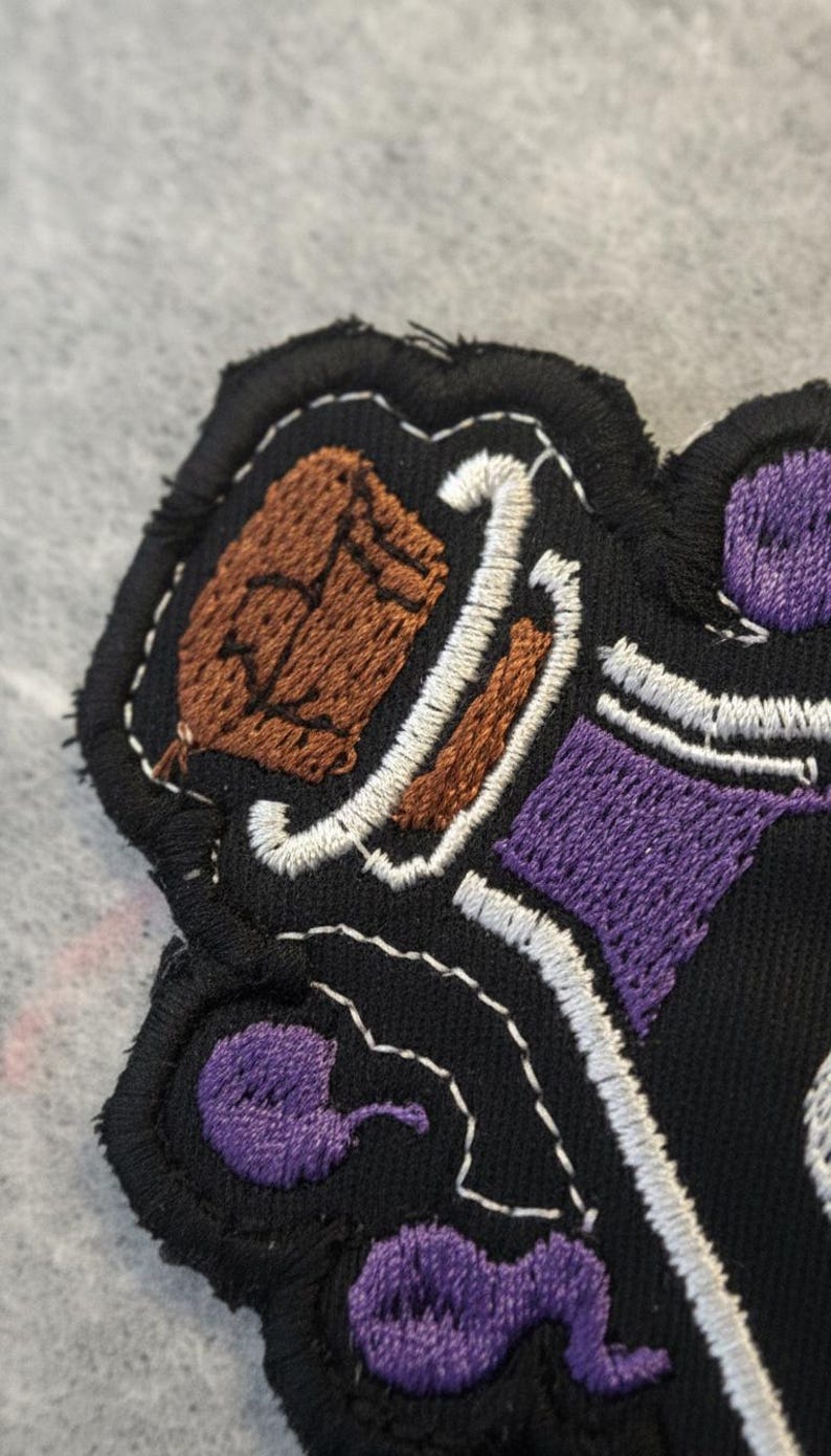 May include: Embroidered patch with a black background and a detailed design featuring brown, purple, and white stitching. The patch has a textured appearance with a stitched border, showcasing a unique, handcrafted aesthetic.