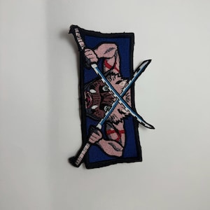 May include: A rectangular embroidered patch featuring a character wearing a boar mask and holding two crossed swords. The design includes a blue background, black border, and detailed stitching in shades of pink, brown, white, and light blue.