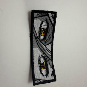 May include: A rectangular embroidered patch featuring a grayscale comic-style illustration of two eyes. The eyes are detailed with white, yellow, and red accents, set against a shaded background, all enclosed within a thick black stitched border.