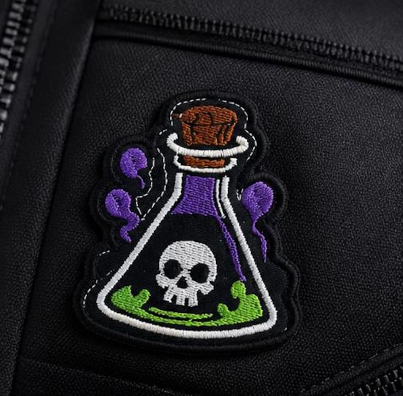 May include: Embroidered patch depicting a cartoon potion bottle with a skull and crossbones. The bottle is purple with a brown cork, and green liquid at the bottom. The patch is on a black background.