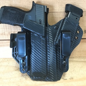 May include: Black carbon fiber pattern holster with a black handgun and magazine. The handgun is marked "SIG Sauer". The holster has two black belt clips and is designed for concealed carry. The background is a light brown wood.
