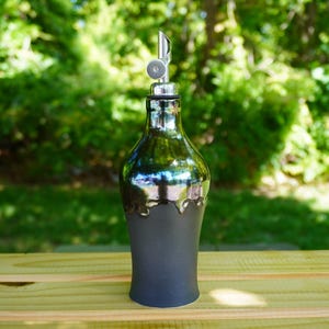 May include: A decorative oil bottle with a silver spout and a dark gray base. The bottle has a metallic, reflective finish on the upper portion, with a unique, dripped design where the metallic finish meets the base. The bottle is on a wooden surface.