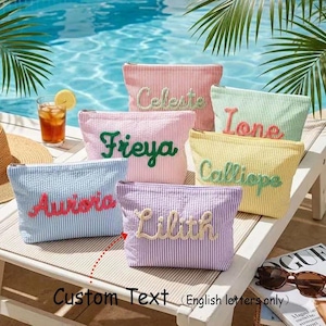 May include: A collection of colorful, striped cosmetic bags with custom text in various colors. The bags are arranged on a white surface near a pool. The bags are in shades of pink, blue, green, yellow, and purple.