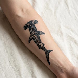 Può includere: Tatuaggio di squalo martello in bianco e nero con un design marmorizzato. Il tatuaggio è sull'avambraccio, mostrando la forma distintiva della testa e del corpo dello squalo. Il design presenta motivi a vortice.