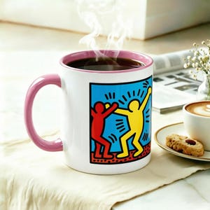 Op de afbeelding: Witte keramische mok met een roze oor en rand, gevuld met donkere koffie en stoom. De mok heeft een kleurrijke afbeelding van twee dansende figuren op een blauwe achtergrond. Een kleine koek en een kopje koffie zijn ook zichtbaar.