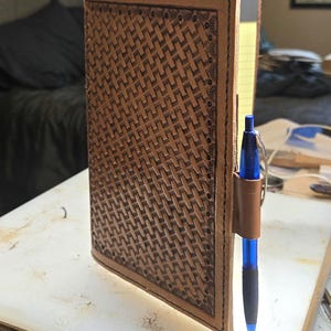 May include: A tan leather notebook cover featuring a stamped basketweave pattern and a stitched border. The cover includes a leather pen loop on the side holding a blue retractable ballpoint pen, with yellow lined paper visible inside the notebook.