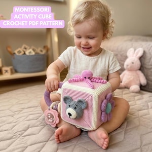 May include: A crochet activity cube in pink, white, and lavender, with a bear, flower, and octopus design. The cube has a handle and a rattle. The image includes the text "Montessori Activity Cube Crochet PDF Pattern".