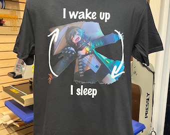 Puede incluir: Una camiseta negra de manga corta con un gráfico de un personaje de estilo anime tumbado con los brazos extendidos. El texto "I wake up" está impreso sobre el personaje y "I sleep" debajo, con flechas blancas rodeando la figura.