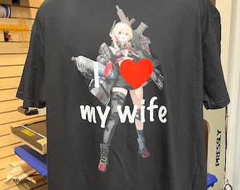Puede incluir: Una camiseta negra de manga corta con un estampado gráfico de un personaje estilo anime que sostiene un rifle y un bote. El diseño incluye un gran símbolo de corazón rojo y el texto blanco "my wife" impreso bajo la ilustración.