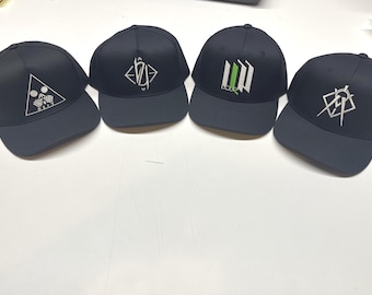 Puede incluir: Cuatro gorras de béisbol azul marino oscuro con viseras planas. Cada gorra presenta un gráfico bordado en blanco único en el panel frontal. Los diseños incluyen un triángulo con texto, una forma de diamante, barras verticales y un compás geométrico.