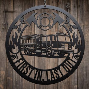 Könnte beinhalten: Schwarzes Metall-Rundschild mit einem Feuerwehrauto, Äxten und einem Malteserkreuz-Emblem. Das Schild enthält den Text "FIRST IN, LAST OUT" und ist zum Aufhängen bestimmt. Der Hintergrund ist eine Holzoberfläche.