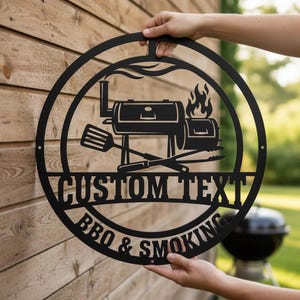 Peut inclure: Panneau noir en métal pour barbecue et fumoir avec un design circulaire. Le panneau présente une illustration de grill, des flammes et les mots "CUSTOM TEXT" et "BBQ & SMOKING". Le panneau est tenu par deux mains.