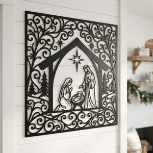 Könnte beinhalten: Ein quadratisches Wandbild aus schwarzem Metall mit einer lasergeschnittenen Krippenszene. Das Design zeigt einen zentralen Stall mit einem Stern darüber, darin Figuren von zwei Erwachsenen und einem Baby in einer Krippe, umgeben von Ranken.