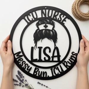 Könnte beinhalten: Schwarze Metall-Wandkunst mit dem Text "ICU Nurse" und "Messy Bun. ICU Runs." In der Mitte befinden sich eine Krankenschwestermütze und ein unordentlicher Dutt, darunter der Name "LISA". Die Kunst ist rund und wird von Händen gehalten.