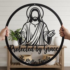 Könnte beinhalten: Schwarze Metall-Wandkunst, die eine Figur mit ausgestreckten Armen zeigt, umgeben von einem Kreis. Der Text "Protected by Grace" und "The Miller Family" befinden sich darunter. Die Kunst wird von zwei Händen gehalten.