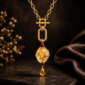 May include: A gold-toned necklace featuring a thick chain with a toggle clasp, a large central oval link, a faceted yellow gemstone, two small dark red beads, and a small teardrop-shaped yellow crystal pendant hanging at the very bottom.
