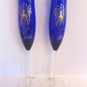 May include: Two cobalt blue glass champagne flutes featuring a gold rim and a gold graphic illustration of a figure surrounded by stars. Each flute has a clear glass stem and a circular base with a small sticker visible on the bottom surface.