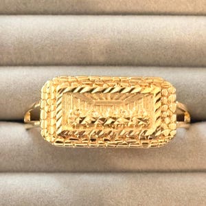 May include: A gold ring featuring a rectangular centerpiece with a textured, pebbled border. The center of the rectangle displays a detailed, engraved scene of the Last Supper, characterized by polished gold surfaces and intricate, reflective facets.