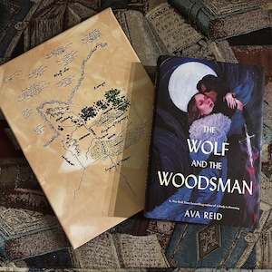 May include: A canvas map with a hand-drawn design and a book titled "The Wolf and the Woodsman" by Ava Reid. The book cover features a couple in a dark, romantic scene under a full moon. The map is tan with black and green details.