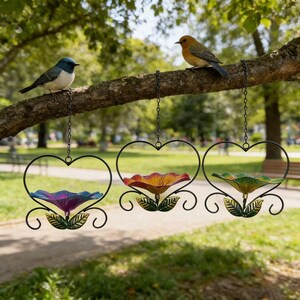 May include: Three hanging heart-shaped bird feeders with colorful flower-shaped bowls. The bowls are purple, red, and green. Two small birds perch on a tree branch above the feeders. The feeders are suspended by chains.