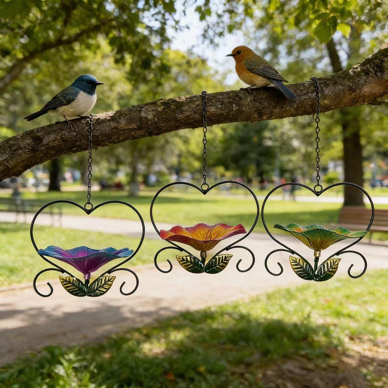 May include: Three hanging heart-shaped bird feeders with colorful flower-shaped bowls. The bowls are purple, red, and green. Two small birds perch on a tree branch above the feeders. The feeders are suspended by chains.