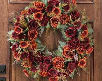 Puede incluir: Una corona circular compuesta por flores artificiales en tonos burdeos intenso, naranja quemado y óxido. El arreglo presenta rosas, dalias y pequeñas flores de relleno intercaladas con follaje verde y tallos finos de aspecto seco.