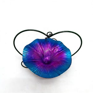 May include: A vibrant, iridescent metal flower sculpture in shades of blue and purple. The flower is attached to a dark green wire heart-shaped frame. The sculpture is set against a white background.