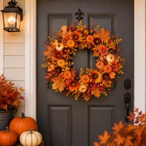 May include: A circular autumn wreath featuring orange and yellow maple leaves, sunflowers, and small pumpkins. The wreath is mounted on a dark gray door, surrounded by a black wall lantern and orange pumpkins on the ground in the foreground.