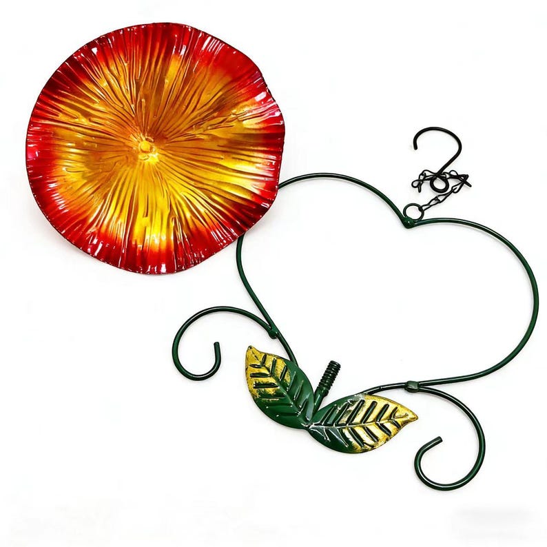 May include: A decorative metal wall art piece featuring a red and yellow flower-shaped dish, a green heart-shaped frame, and green and gold leaves. The piece includes a chain and hook for hanging.