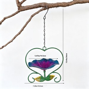 May include: A hanging bird feeder shaped like a heart, with a purple and blue flower-shaped bowl. The feeder is 13.4 inches tall and 7.28 inches wide, with a 5.47-inch bowl. It is suspended by a chain and hook from a tree branch.