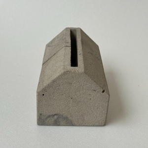 May include: A small, house-shaped concrete object in a light grey colour. The object has a long, narrow slot on the top and two small holes on the front. The surface has a slightly rough texture.