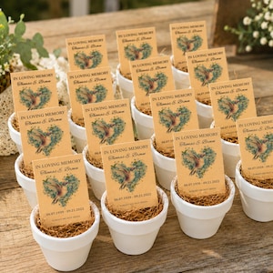 May include: A collection of small white ceramic pots, each containing brown fiber and a rectangular tan card. The cards feature the text IN LOVING MEMORY, Thomas S. Brown, 06.07.1939 - 09.21.2022, and a watercolor illustration of a hummingbird inside a green heart.