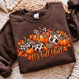 May include: A dark brown crewneck sweatshirt featuring a graphic of multiple pumpkins with various patterns including buffalo plaid, leopard print, and cow print, accented by orange autumn leaves and the text It's Fall Y'all.