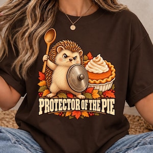Peut inclure: Un t-shirt marron foncé avec un motif représentant un hérisson tenant une cuillère en bois et un bouclier en couvercle métallique, à côté d'une tarte à la citrouille. Le design inclut des feuilles d'automne et le texte "PROTECTOR OF THE PIE".