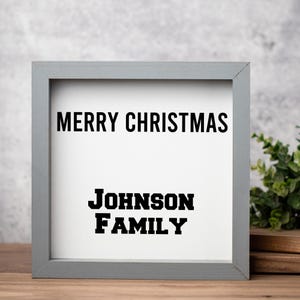 May include: A framed sign with a gray border and a white interior. The words "MERRY CHRISTMAS" are printed in bold black letters at the top, with "JOHNSON FAMILY" below. The sign is on a wooden surface.