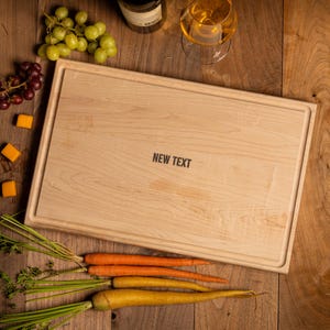 May include: A rectangular wooden cutting board with a grooved border and the text NEW TEXT engraved in the center. The board is surrounded by green grapes, orange carrots, yellow carrots, and two small yellow cubes on a wooden surface.