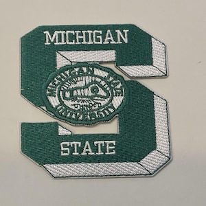 May include: A green and white embroidered patch in the shape of the letter "S". The patch features the words "MICHIGAN" and "STATE" in white lettering. A circular emblem in the center reads "MICHIGAN STATE UNIVERSITY".