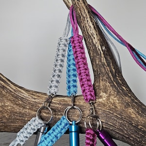 May include: A collection of colorful paracord keychains with small, cylindrical containers. The keychains are in shades of silver, blue, and magenta, and are attached to metal rings. The containers are silver, blue, and purple. The items are displayed against a natural wood background.