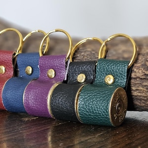 May include: Five leather keyrings in red, blue, purple, black, and green. Each keyring has a gold-coloured ring and a brass button. The base of each keyring is a brass bullet casing.