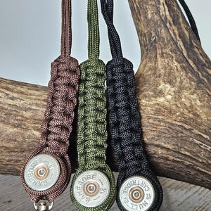May include: Three shotgun shell keychains in brown, olive green, and black, each with a braided cord and a silver-tone clasp. The shell casings have the text "GAME BORE" and "HULL 12". The keychains are displayed against a natural antler.