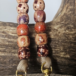 May include: Two beaded lanyards with gold-colored clasps. Each lanyard features a series of wooden beads in shades of brown, orange, and purple, with intricate patterns. The lanyards are attached to a beige cord.