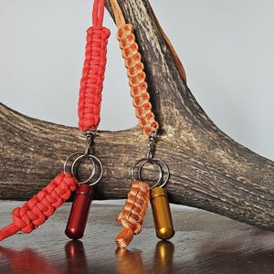 May include: Two keychains with braided cords in red and orange, each attached to a small, cylindrical container. The keychains hang in front of a large, textured antler, creating a rustic and natural aesthetic. The containers are metallic.